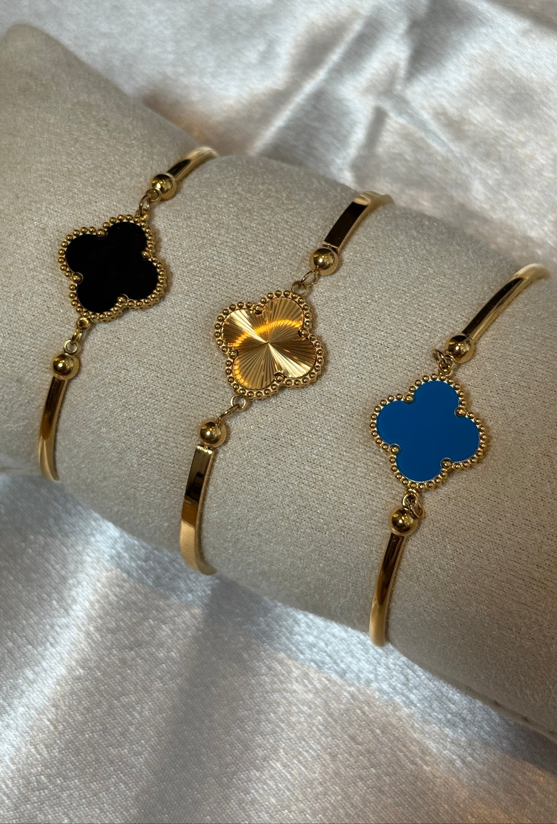 Drei goldene Armreifen mit kleeblattförmigen Anhängern in Schwarz, Gold und Blau – elegant präsentiert auf einem cremefarbenen Samtkissen vor glänzendem Satin-Hintergrund.