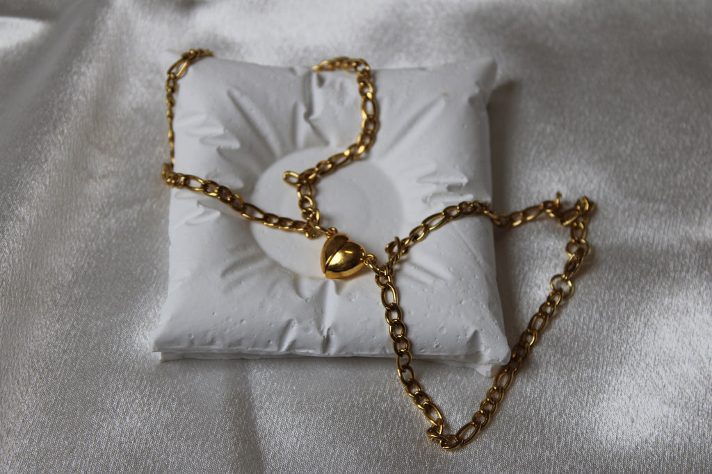 Zwei goldfarbene Gliederarmbänder mit herzförmigen Verschlüssen, dekorativ platziert auf einem weißen Schmuckkissen vor glänzendem Satin-Hintergrund.