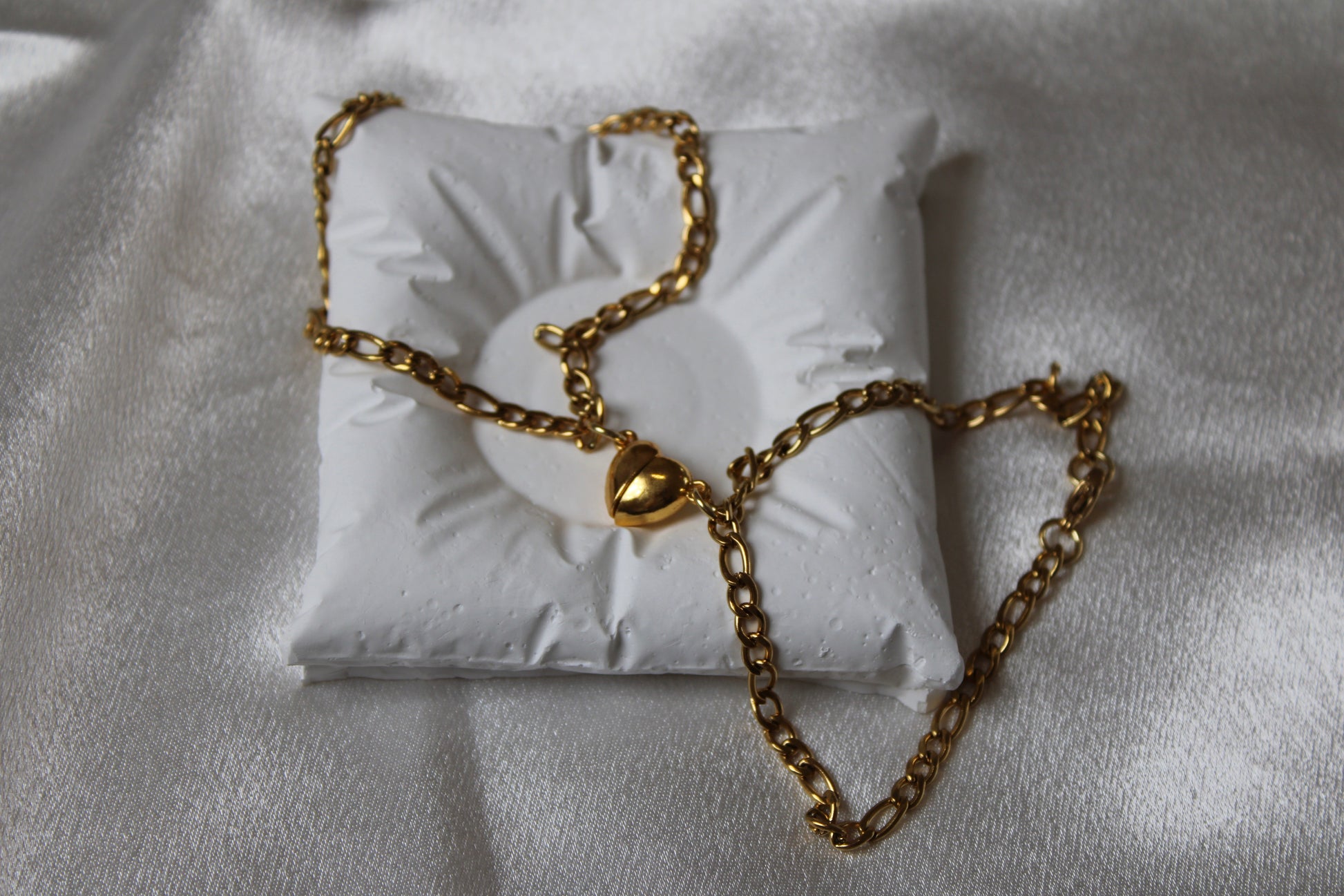 Zwei goldfarbene Gliederarmbänder mit herzförmigen Verschlüssen, dekorativ platziert auf einem weißen Schmuckkissen vor glänzendem Satin-Hintergrund.