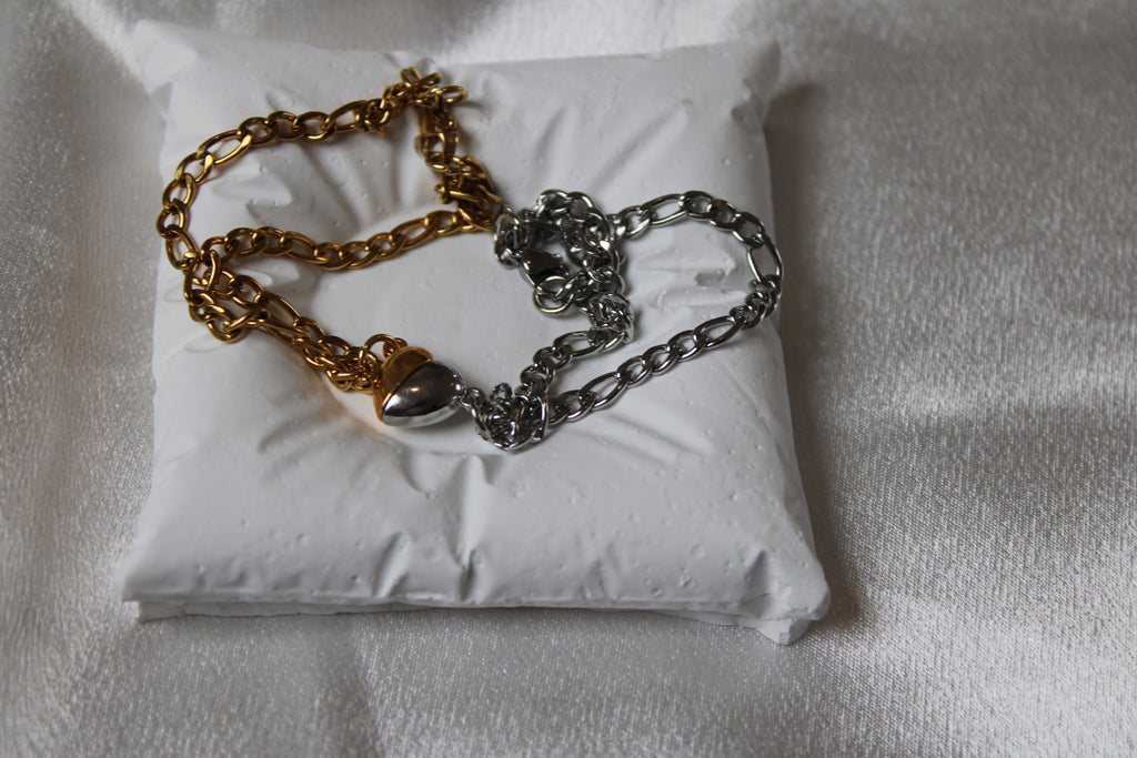 Zweifarbige Gliederarmbänder – eines in Gold, das andere in Silber – mit herzförmigen Verschlüssen, dekorativ platziert auf einem weißen Schmuckkissen vor glänzendem Satin-Hintergrund.