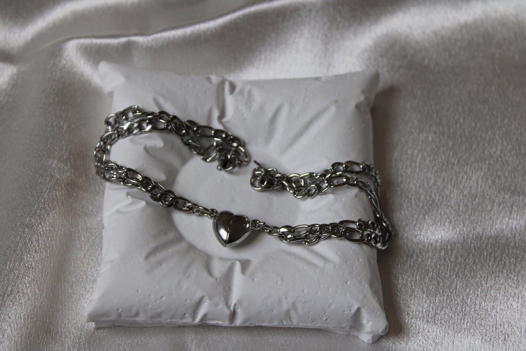 Zwei silberfarbene Gliederarmbänder mit herzförmigen Verschlüssen, dekorativ platziert auf einem weißen Schmuckkissen vor glänzendem Satin-Hintergrund.