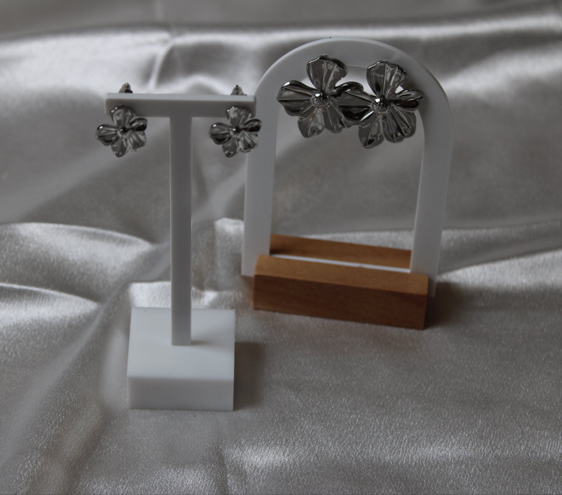 Zwei Paar silberfarbene Blütenohrringe – ein großes und ein kleines Modell – elegant präsentiert auf weißen und hölzernen Schmuckständern vor schimmerndem Satin-Hintergrund.