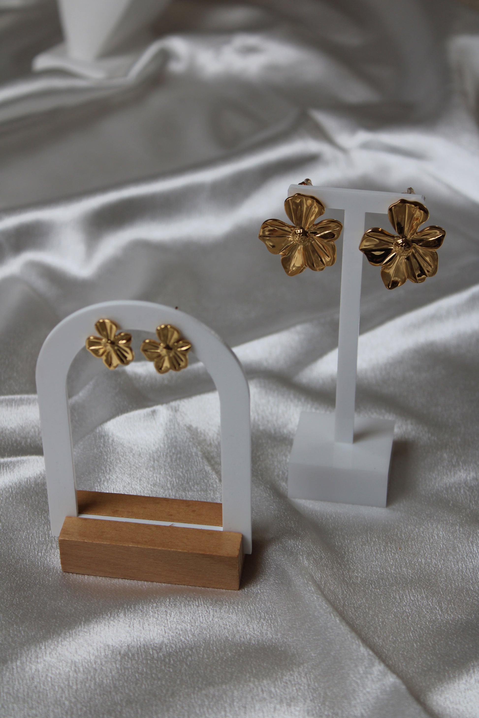 Zwei Paar goldfarbene Blütenohrringe – ein großes und ein kleines Modell – elegant präsentiert auf weißen und hölzernen Schmuckständern vor schimmerndem Satin-Hintergrund.