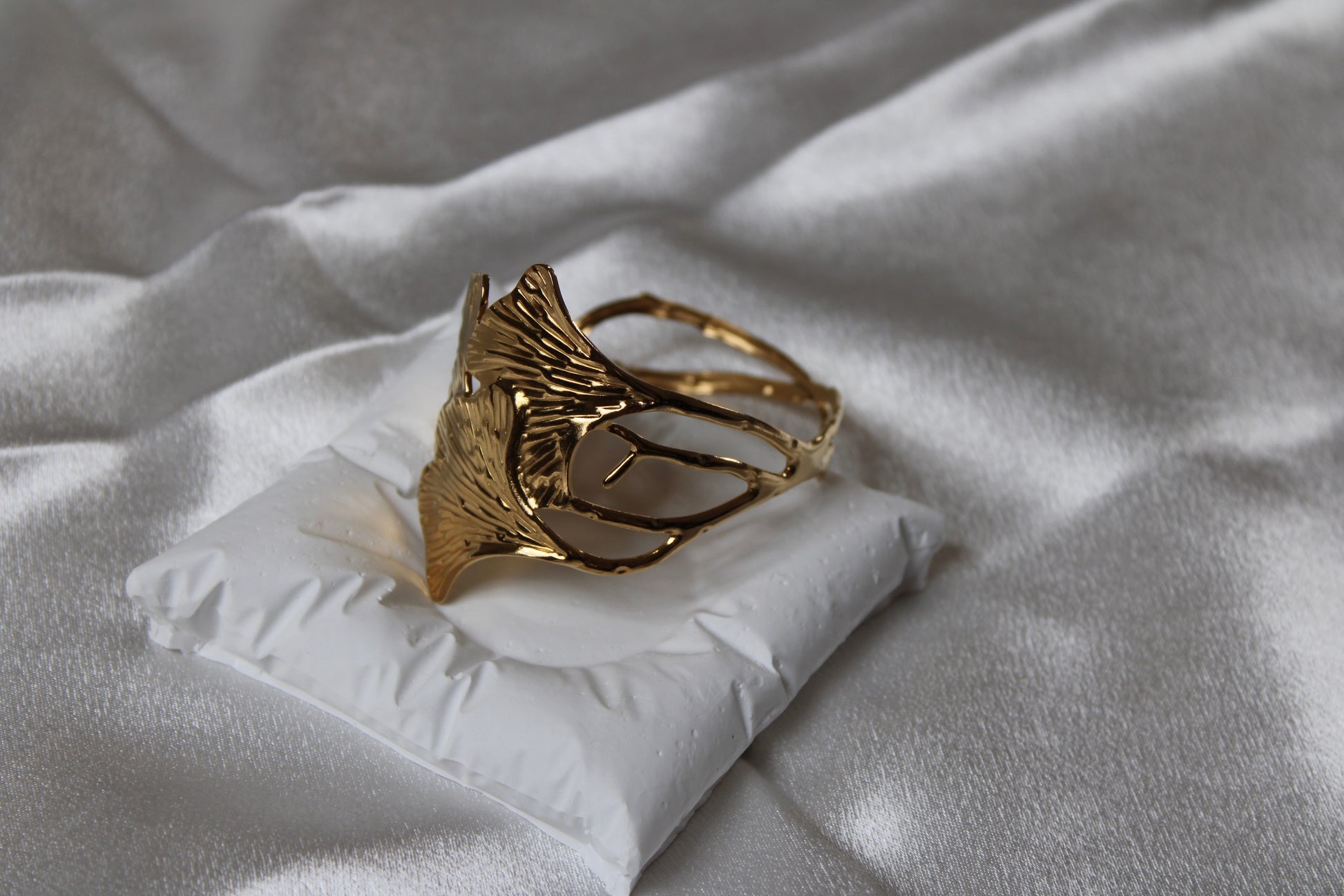 Goldfarbener, offener Armreif in floraler Blattform mit strukturierter Oberfläche – edel drapiert auf weißem Schmuckkissen mit Satin-Hintergrund.