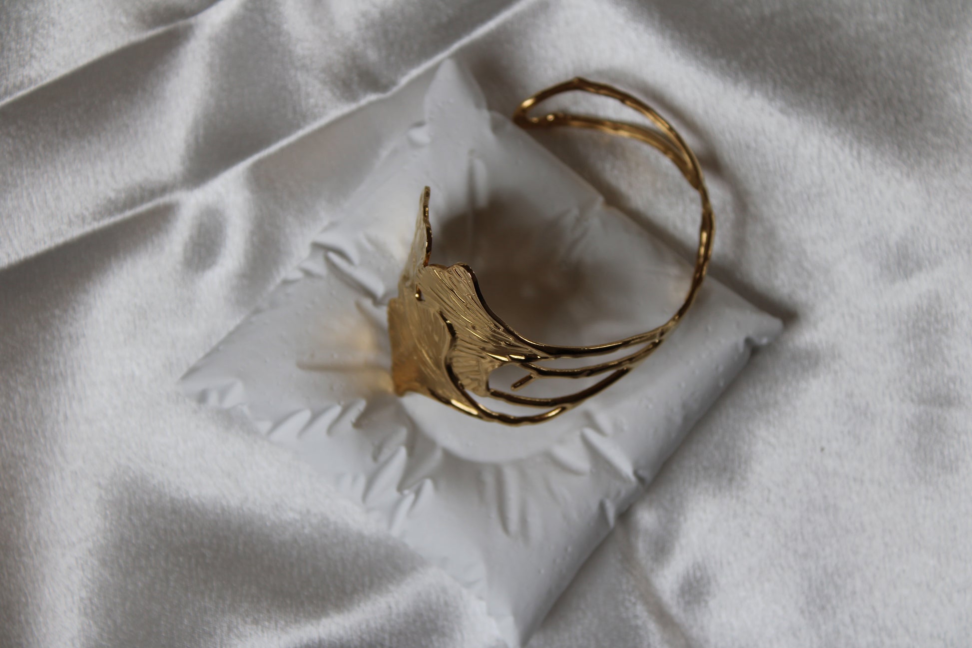 Goldfarbener, offener Armreif in floraler Blattform mit strukturierter Oberfläche – edel drapiert auf weißem Schmuckkissen mit Satin-Hintergrund.