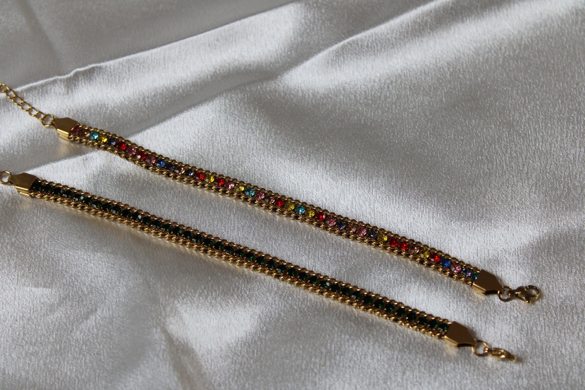 Zwei filigrane Armbänder auf weiß glänzendem Stoff drapiert. Das obere mit bunten Kristallen, das untere mit grünen Kristallen. 