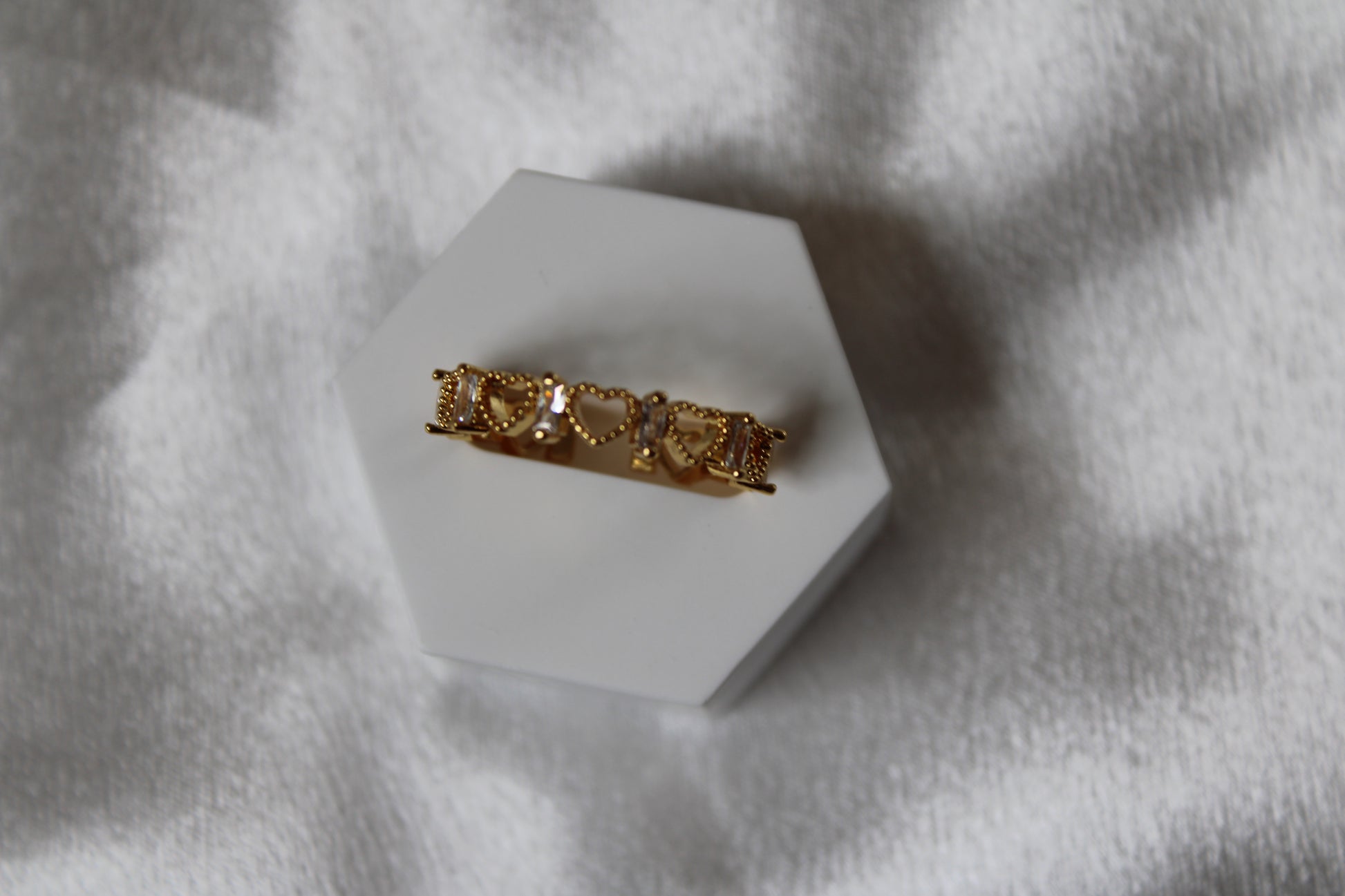 Goldfarbener Ring mit durchgehenden kleinen Herzformen, verziert mit funkelnden weißen Steinen, auf weißem Schmuckständer präsentiert.